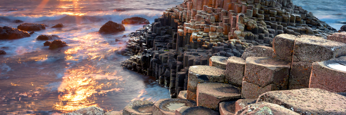 The giants causeway at sunset, the sun is just above the sea on the horizon and glints on the surf of the waves lapping at the rocks.